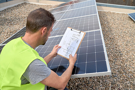 Ein Photovoltaik-Experte prüft eine Solaranlage auf einem Flachdach und hält dabei einen Plan in der Hand. 