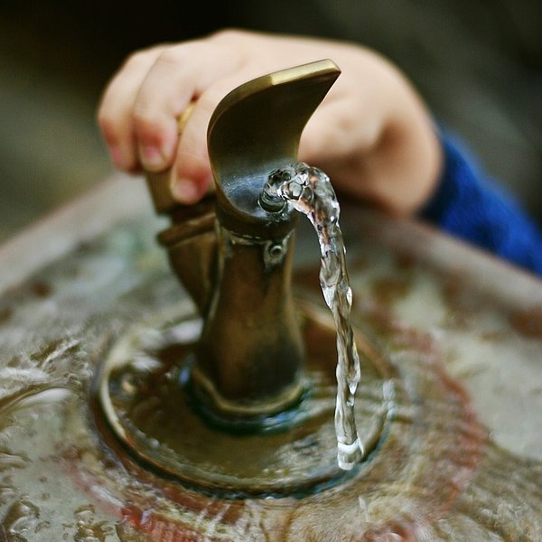 Eine Kinderhand bedient einen Trinkbrunnen aus dem frisches Wasser fließt.