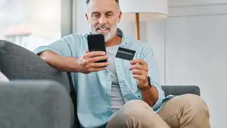 Mann sitzt auf Sofa mit Handy und Kreditkarte in der Hand