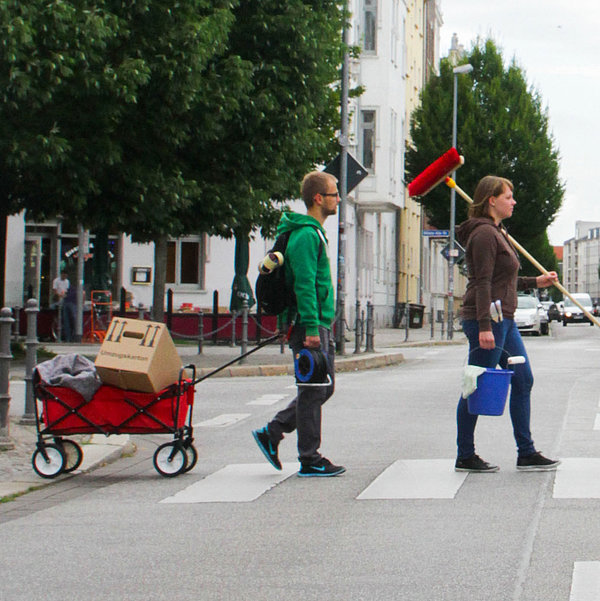 Vier junge Leute laufen mit Besen, Maler- und Putzsachen und Bollerwagen über einen Zebrastreifen in Magdeburg Stadtfeld.