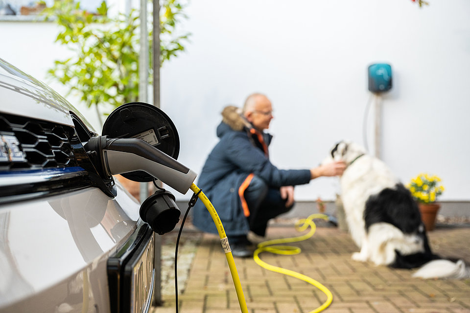 Zeit für die wichtigen Dinge. Ihr E-Auto lädt mit der SWM Wallbox bequem zu Hause. Ein Elektroauto lädt, der Stecker steckt in der Ladebuchse. Im Hintergrund streichelt ein Mann seinen Hund, ebenso ist die SMW Wallbox an der Hauswand zu erkennen.