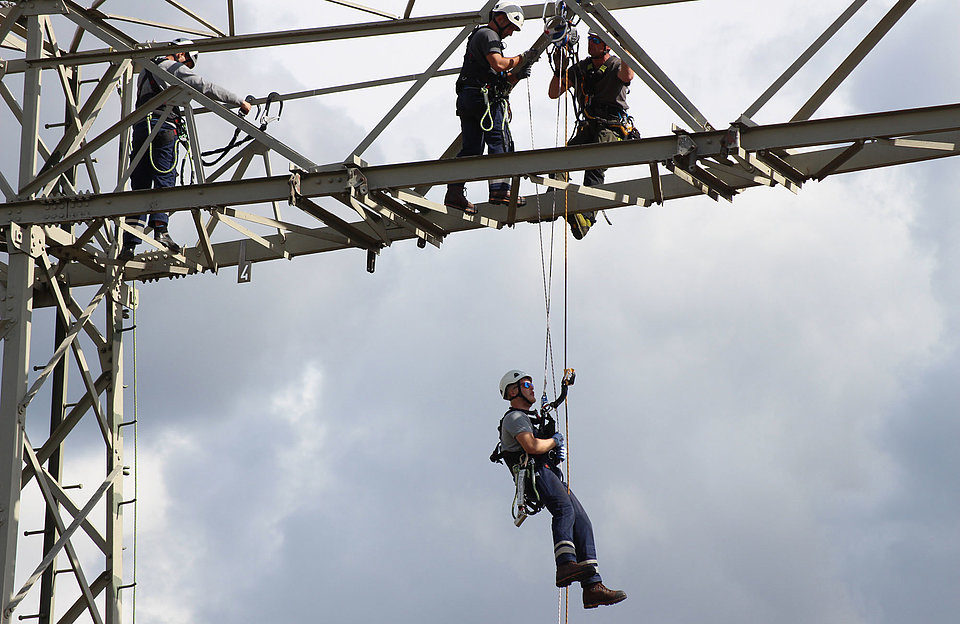 Ein SWM Mitarbeiter seilt sich vom Hochspannungsmast ab. Zwei Mitarbeiter stehen noch oben auf dem Mast.