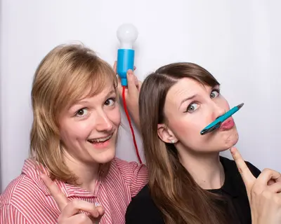Zwei Mitarbeiterinnen von SWM blicken in die Kamera mit Kugelschreiber und Glühlampe als Accessoires