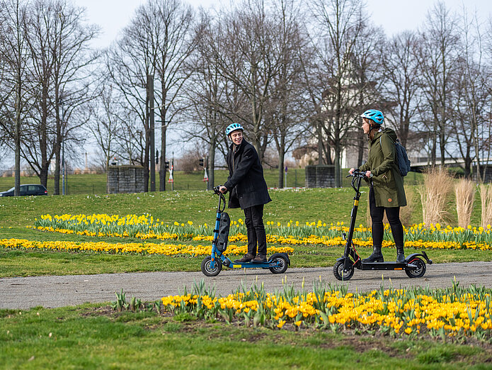 Ein Mann und eine Frau fahren auf dem E-Scooter im Frühling durch den Elbauenpark in Magdeburg. 