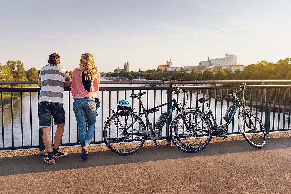Beim E-Bike Ausflug gemeinsam die Aussicht auf die Stadt genießen. Zwei Personen stehen auf der Jerusalembrücke in Magdeburg, schauen auf die Elbe im Hintergrund sind der Dom und die Johanniskirche zu sehen. Neben Ihnen stehen die SWM E-Bikes.