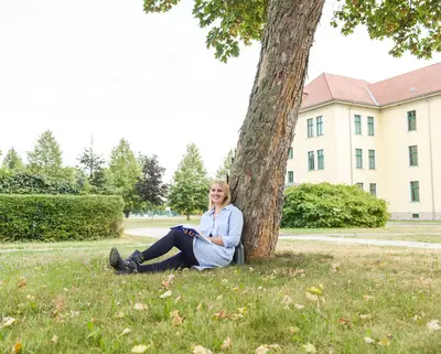 Studentin der dualen Ausbildung bei SWM sitzt auf dem Hochschul-Campus am Baum und liest