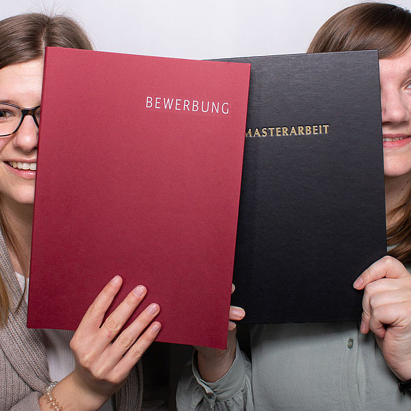 Zwei Mitarbeiterinnen von SWM zeigen eine Bewerbung und eine Masterarbeit in die Kamera
