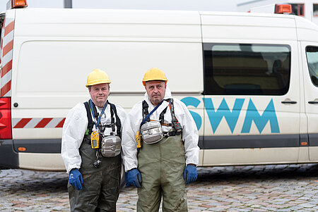 zwei SWM Mitarbeiter der Abwasserentsorgung stehen in Arbeitskleidung vor einem Firmenfahrzeug