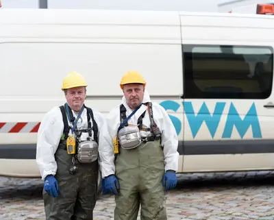 zwei SWM Mitarbeiter der Abwasserentsorgung stehen in Arbeitskleidung vor einem Firmenfahrzeug