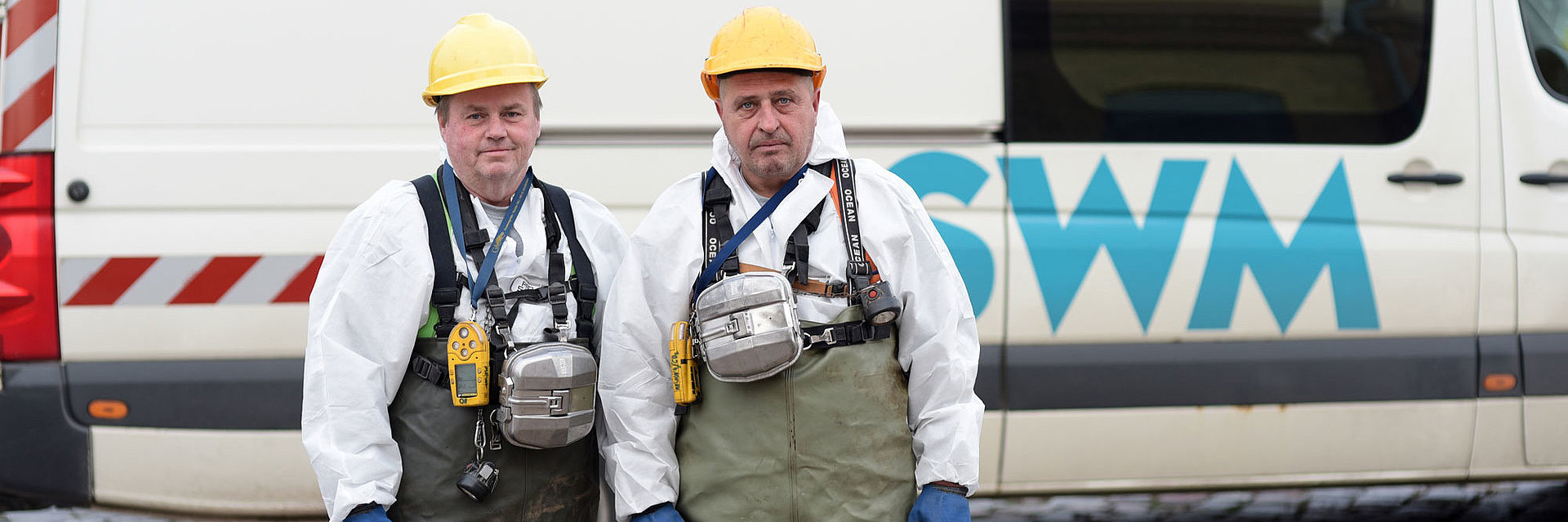 zwei SWM Mitarbeiter der Abwasserentsorgung stehen in Arbeitskleidung vor einem Firmenfahrzeug