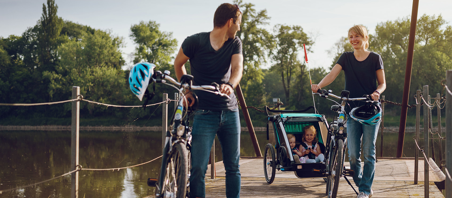 Eine Familie schiebt im Sommer SWM E-Bikes über eine Brücke im Stadtpark. Die Kinder sitzen im Fahrradanhänger. 
