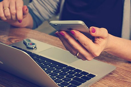 Eine Frau sitzt am Laptop und schaut auf Ihr Smartphone.