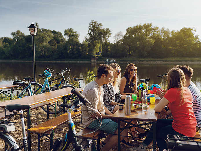 Sechs junge Menschen sitzen im Biegarten am Mückenwirt in Magdeburg an der Elbe. Im Hintergrund sind die SWM E-Bikes zu sehen. 