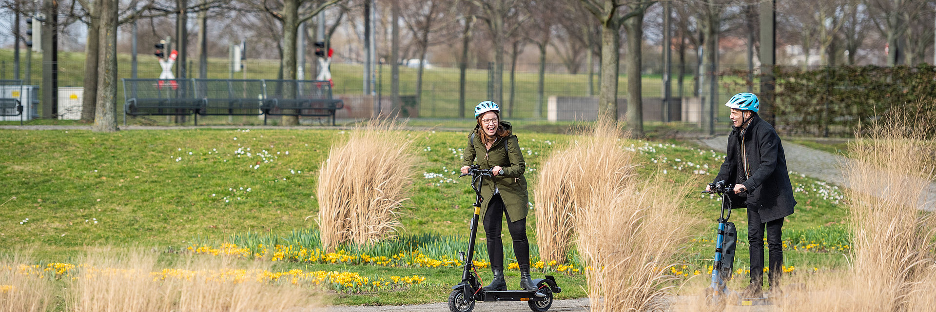 Eine Frau und ein Mann fahren durch den Elbauenpark in Magdeburg mit einem SWM E-Scooter. Im HIntergrund ist der Jahrtausendturm zu sehen.