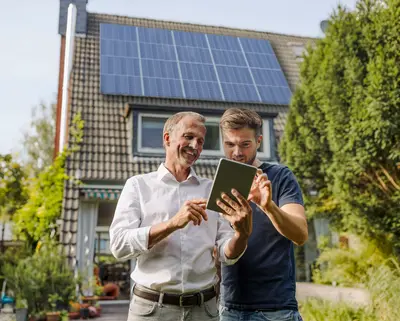 Zwei Männer stehen vor einem Haus mit PV Anlage und schauen auf ein Tablet