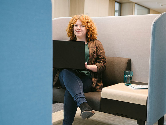 Auszubildende sitzt mit Laptop auf dem Schoß in einer Besprechungsinsel und lacht