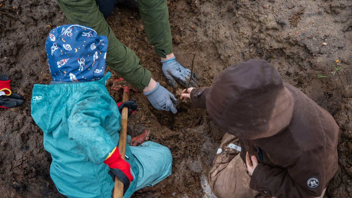 Zwei Kinder sitzen auf dem Boden und pflanzen mit Hilfe einer erwachsenen Person einen Baum