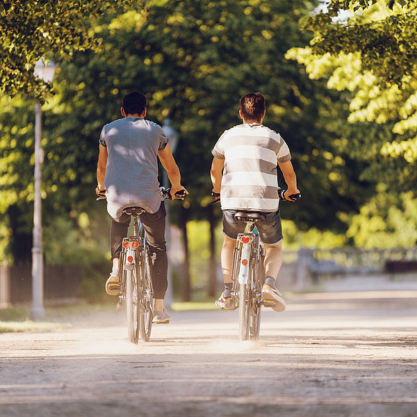 Zwei Männer fahren auf SWM E-Bikes im Sommer durch eine Allee in Magdeburg