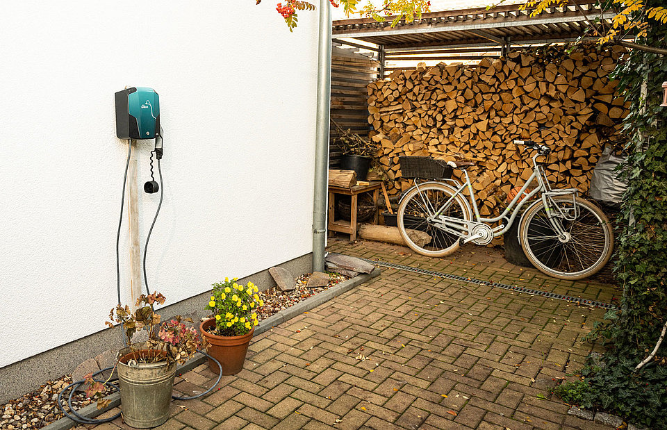 SWM Wallbox an einer Hauswand mit Holzstapel und Fahrrad im Hintergrund