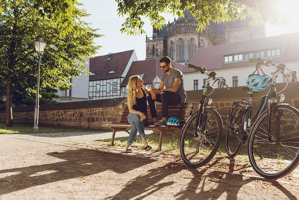 Ein Pärchen sitzt bei Sonnenschein auf einer Bank am Fürstenwall in Magdeburg, neben Ihnen stehen zwei SWM-E-Bikes. 