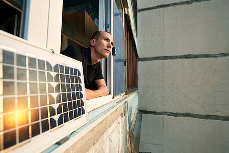 Mann lehnt sich aus einem Fenster, daneben befindets ich eine Mini-PV-Anlage