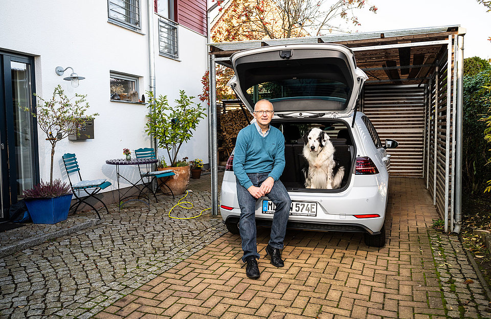 E-Laden zu Hause Mann und Hund sitzen im offenen Kofferraum vor ihrem Haus während ihr E-Auto mit der SWM Wallbox lädt