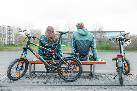 Zwei Leute sitzen auf einer Bank und daneben stehen zwei Räder "Emil" von trittfest