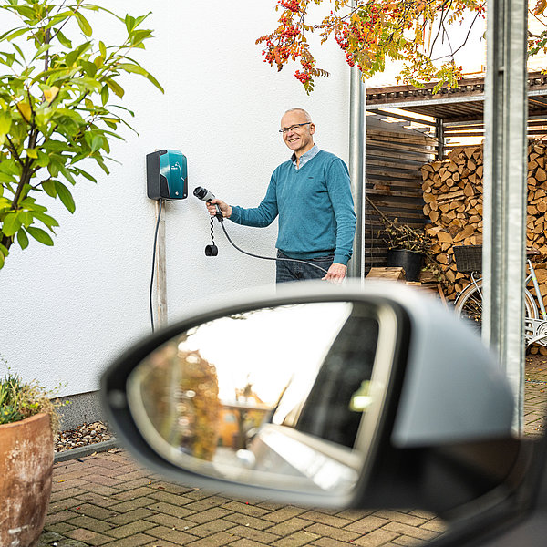 Ein Mann hält den Stecker eines Elektroautos in Richtung einer Wallbox zum Laden des Autos von zu Hause. Die Wallbox ist an einer Hauswand befestigt.