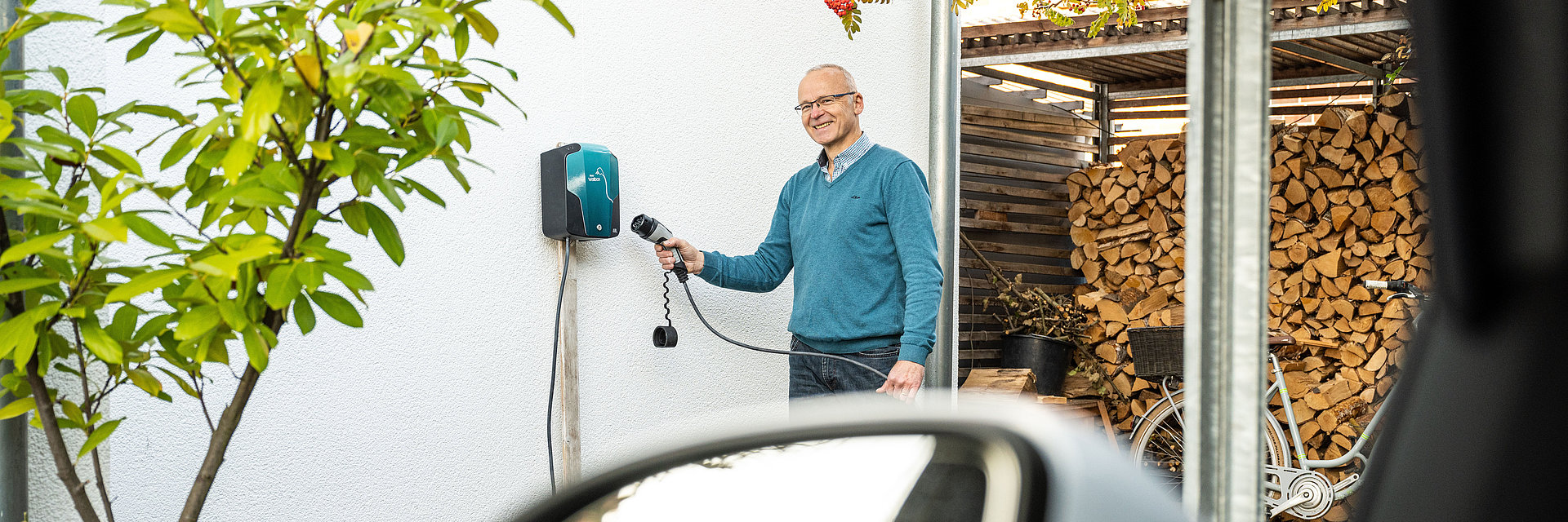 Ein Mann hält den Stecker eines Elektroautos in Richtung einer Wallbox zum Laden des Autos von zu Hause. Die Wallbox ist an einer Hauswand befestigt.