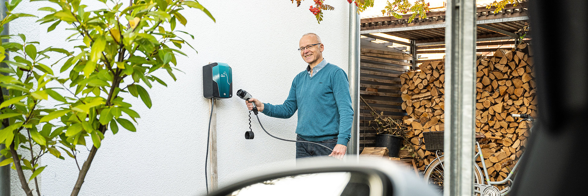 Ein Mann hält den Stecker eines Elektroautos in Richtung einer Wallbox zum Laden des Autos von zu Hause. Die Wallbox ist an einer Hauswand befestigt.