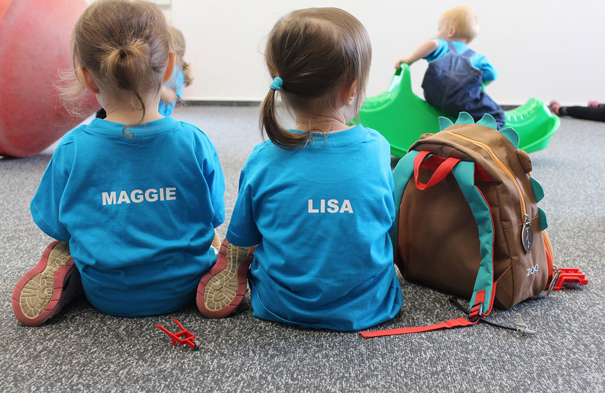 SWM Kita Kinder Zwei Kinder im SWM T-Shirt sitzen mit dem Rücken zur Kamera in der SWM Kita