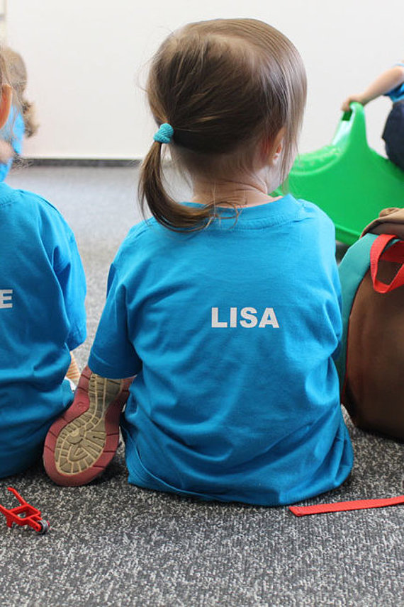 Zwei Kinder im SWM T-Shirt sitzen mit dem Rücken zur Kamera in der SWM Kita