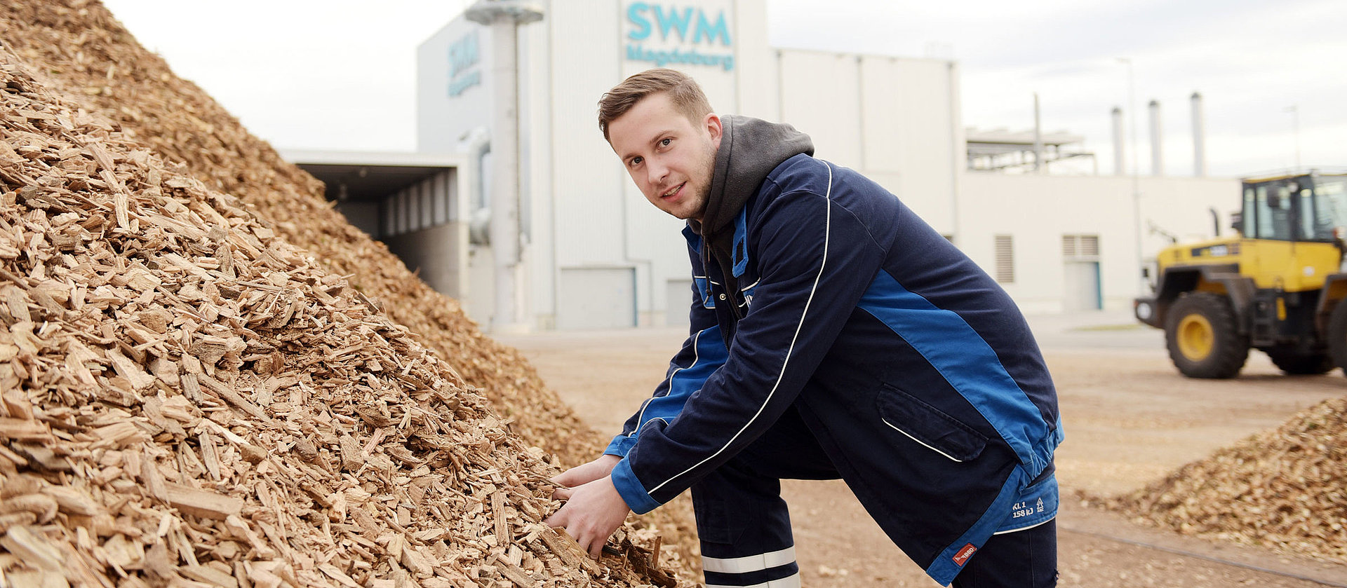 SWM Mitarbeiter am Biomasseheizkraftwerk SWM Mitarbeiter greift in einen großen Berg Holzhackschnitzel vor dem SWM Biomasseheizkraftwerk.