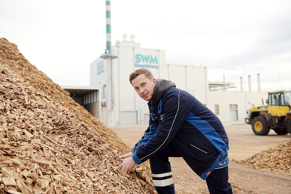 SWM Mitarbeiter greift in einen großen Berg Holzhackschnitzel vor dem SWM Biomasseheizkraftwerk.