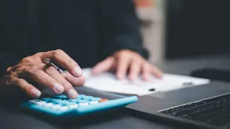 Mensch sitzt vor Laptop und tippt etwas in einen Taschenrechner ein