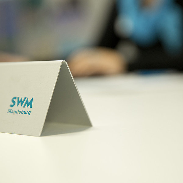 Es ist ein Namenschild der SWM Kundenberatung zu sehen. Im Hintergrund sitzt ein Mitarbeiter des Kundenserviceteams.