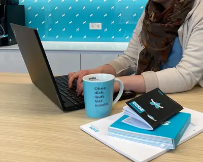 Frau sitzt am Laptop mit SWM Tasse, die eine Aufschrift "Ohne dich läuft hier nüscht" hat